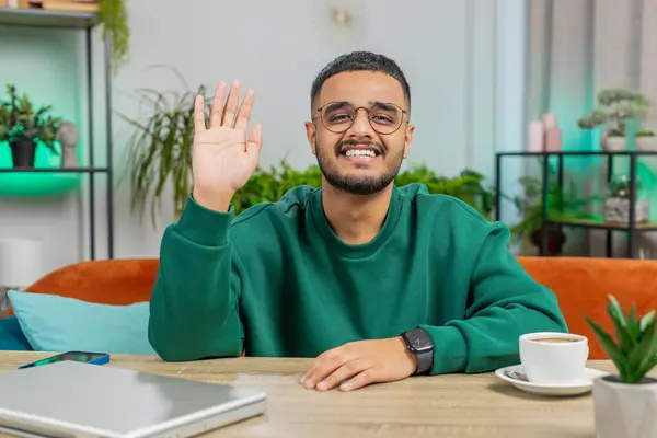 Hello. Indian young man smiling friendly at camera and waving hands gesturing hello hi, greeting or goodbye welcoming with hospitable expression at home office. Arabian guy sitting at table in room