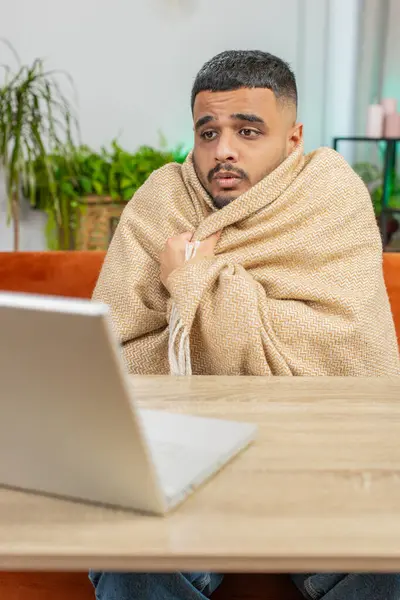 Sick ill Indian man freelancer working on laptop wrapped in plaid sit shivering from cold drinking hot coffee in unheated home office without heating due debt. Unhealthy Arabian guy try to warming up