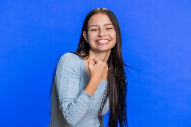 Başarılı zaferi kutlayan neşeli bir kadın, doğum gününü kazanan, piyango vurgunu hedefine ulaşan bir kadın iyi haber, olumlu haber, zafer. Mavi stüdyo duvarında gülümseyen esmer kız.