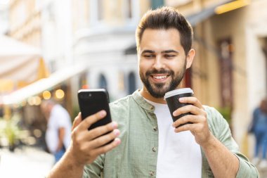 Mutlu gülümseyen adam akıllı telefon kullanıyor mesajlar yazıyor internet sosyal medya uygulamalarına göz atıyor internette sohbet ediyor sabahın tadını çıkarıyor sıcak kahve içmek için dışarı çıkıyor. Şehir caddesinde yürüyen bir adam.