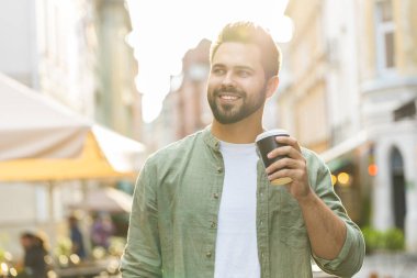 Mutlu sakallı genç adam sabah kahvesinin tadını çıkarıyor sıcak içecek ve dışarıda gülümsüyor. Dinleniyorum, ara veriyorum. Şehirdeki Sunshine Caddesi 'nde yürüyen ve kahve içen bir adam. Dışarıdaki kasaba yaşam tarzları