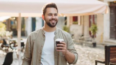 Mutlu sakallı genç adam sabah kahvesinin tadını çıkarıyor sıcak içecek ve dışarıda gülümsüyor. Dinleniyorum, ara veriyorum. Şehirdeki Sunshine Caddesi 'nde yürüyen ve kahve içen bir adam. Dışarıdaki kasaba yaşam tarzları