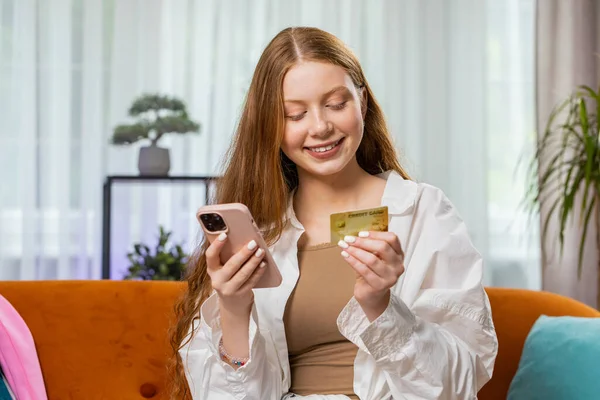 Teenager girl using credit bank card and smartphone while transferring money, purchases online shopping cashless, order food delivery at home apartment indoors. Happy child sitting in room on couch