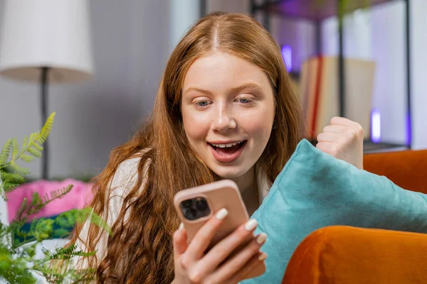 Happy teenager girl use mobile smartphone typing browsing say Wow yes found out great big win good news celebrate lottery jackpot doing winner gesture. Portrait of child at home in room lying on sofa