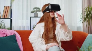 Genç kızıl saçlı kız sanal gerçeklik teknolojisi VR uygulama kulaklığı kullanarak 3D 360 video oyunu simülasyonu yapıyor ve modern ev dairesinde film izliyor. Gözlüklü çocuk kanepeye oturur.
