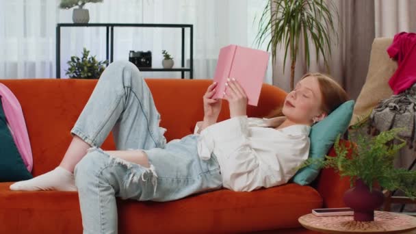 十几岁的红头发女孩轻松地阅读有趣的书籍 翻开书页 享受着文学 在舒适的沙发上休息 快乐的孩子躺在家里的客厅里沙发上 — 图库视频影像