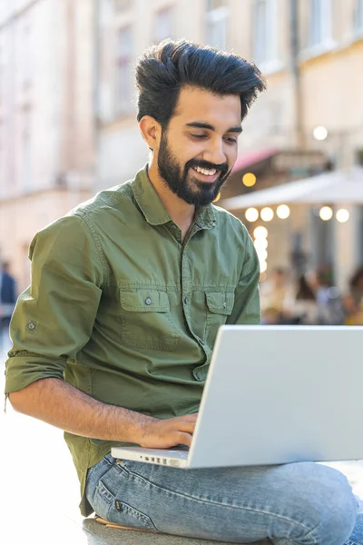 Indian man freelancer working online distant job with laptop sits in city street browsing website chatting outdoor during break. Guy looking at notebook screen sends messages watching movies. Vertical