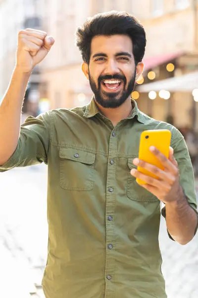 Indian man use mobile smartphone celebrating win good message news, lottery jackpot victory, giveaway online outdoors. Happy adult guy tourist walking in urban city street. Town lifestyles. Vertical