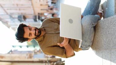 Şehir sokağında oturan Hintli serbest çalışan, laptop kamerasına bakan, arkadaşlarıyla, ailesiyle ya da müşterisiyle görüntülü konuşma yapan, uzak sohbetlerden hoşlanan. El sallayan adam. Dikey