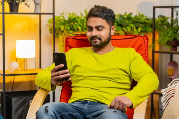 Indian man sits on chair uses mobile phone smiles at home room apartment. Hindu Arabian guy texting share messages content on smartphone social media applications online watching relax movie video