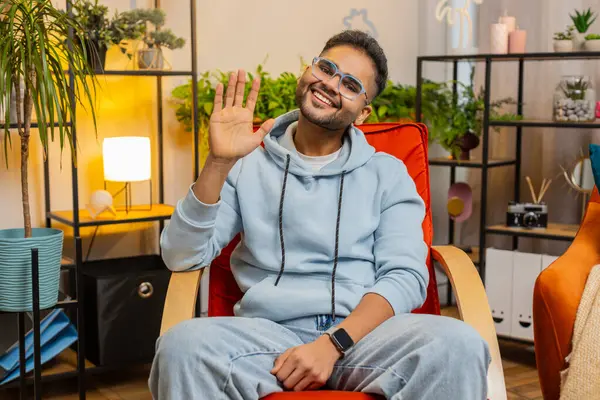 Hello. Indian handsome man smiling friendly at camera and waving hands gesturing hello hi, greeting or goodbye welcoming with hospitable expression at home. Arabian guy sitting on chair in living room