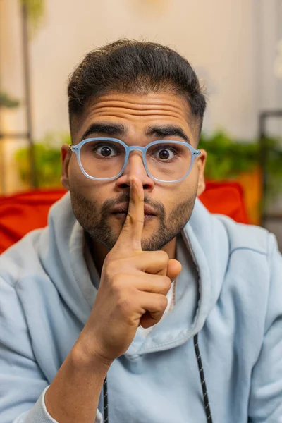 Shh be quiet please. Indian young man presses index finger to lips makes silence hush gesture sign do not tells gossip secret. Arabian guy at home apartment living room sitting on chair. Vertical