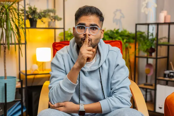 Shh be quiet please. Middle eastern man presses index finger to lips makes silence hush gesture sign do not tells gossip secret. Bearded arabian guy at home apartment living room sitting on chair
