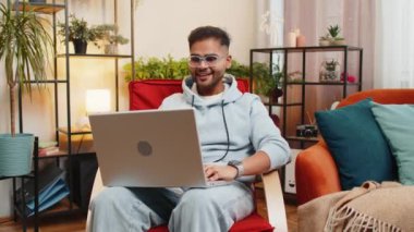 Şaşırmış Hintli genç adam dizüstü bilgisayar kullanıyor, iyi haber mesajı alıyor ani zaferin şokuyla piyango kazanma ikramiyesini kutluyor evdeki evdeki dairede sandalyede oyun oynuyor.