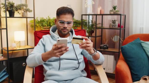 印度男子在汇款时使用信用卡和智能手机 在家里购买网上购物时没有现金 在家里购买食品 快乐的阿拉伯人坐在椅子上的房间里 — 图库视频影像