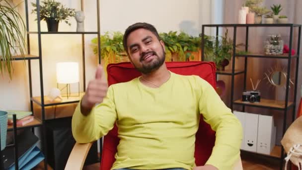 同じように ハッピーエキサイティングなインド人男性は 良いニュースの肯定的なフィードバックをサインのように親指を示すカメラで承認して見ています 若いアラビア人男性が自宅のリビングルームで椅子に座っています — ストック動画