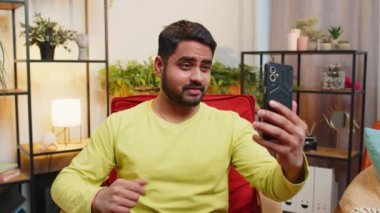 Mutlu Hintli blog yazarı akıllı telefondan selfie çekiyor, abonelerle online video görüşmesi yapıyor, sosyal medya için hikayeler kaydediyor. Modern apartman dairesindeki adam sandalyede oturuyor.