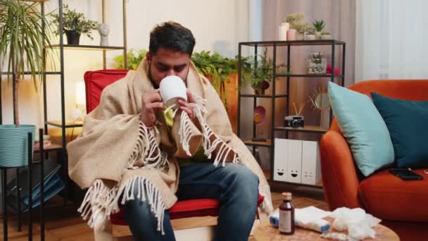 身穿格子呢衣服的生病的印度人孤身一人坐在沙发上 在没有暖气的公寓里喝着热茶 吓得直哆嗦 没有欠债 不健康的阿拉伯人坐在椅子上 觉得不舒服 试着热身 — 图库视频影像