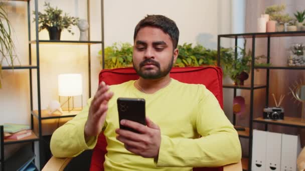不満なアラビア人の男性は スマートフォンのタイピングブラウジングを使用し 突然の宝くじのゲームの結果 悪いニュース 財産の損失 ウイルスに驚くようになります 椅子の自宅の部屋にいる若いインド人男性 — ストック動画