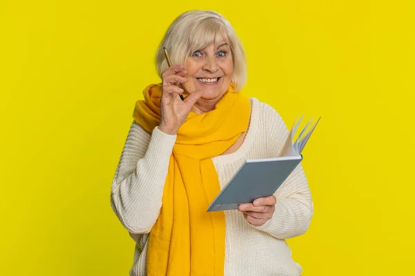 Thoughtful journalist old senior woman making notes, writing down thoughts with pen into notepad notebook diary, to do list, good idea inspiration. Elderly grandmother isolated on yellow background