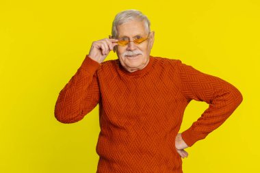 Turuncu güneş gözlüğü takan baştan çıkarıcı, stil sahibi yaşlı bir adamın portresi, büyüleyici bir gülümseme, zafer kutlamaları, olumlu haberler. Mutlu yaşlı büyükbaba emeklisi sarı arka planda