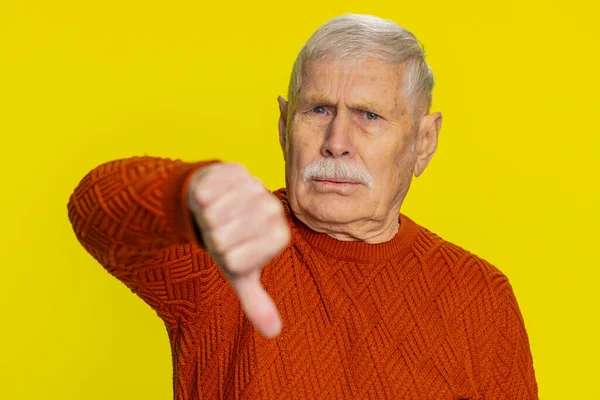 Dislike. Upset unhappy senior old man showing thumbs down sign gesture, expressing discontent, disapproval, dissatisfied negative feedback. Mature pensioner grandfather isolated on yellow background