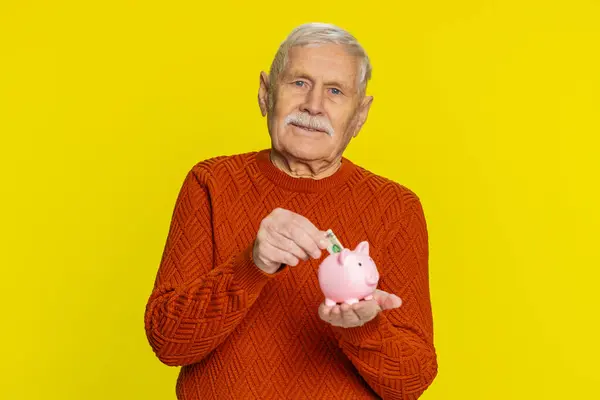 Senior rich old man putting dollar pocket money cash into piggy bank saving for future needs. Grandfather pensioner with coins box finance income growth salary pension, economy on yellow background