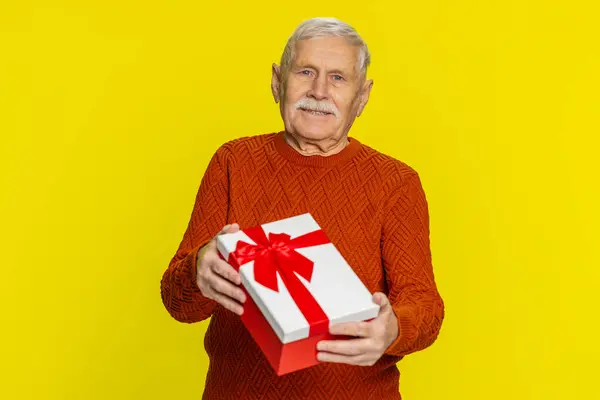 Positive smiling senior man presenting birthday gift box stretches out hands, offer wrapped present career bonus, celebrating party, promotion discount sale. Elderly grandfather on yellow background