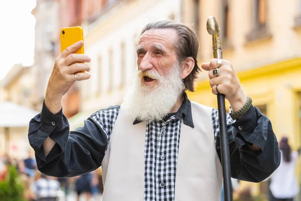 Senior elderly man use mobile smartphone celebrating win good message news, lottery jackpot victory, giveaway online outdoors. Happy old grandfather tourist walking in city street. Town lifestyles