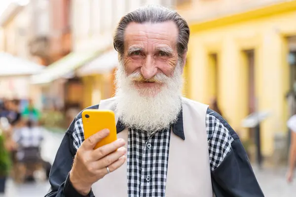 Happy smiling bearded senior old man use smartphone typing text messages browsing internet social media web app working chatting online. Mature grandfather tourist looking at camera in city street
