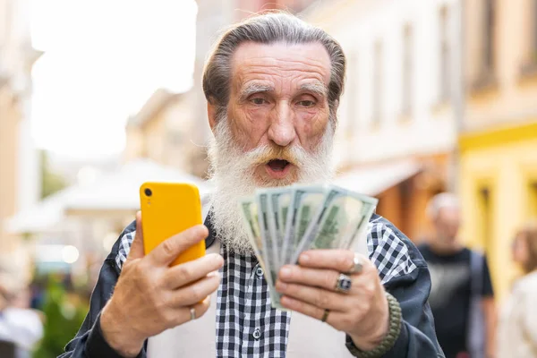 Happy rich senior old man counting holding money dollar cash, use smartphone calculator app, plans to order gifts, food delivery online, booking hotel room. Elderly grandfather tourist in city street