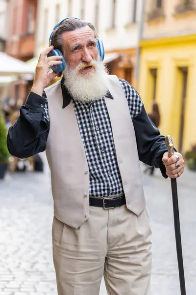 Happy relaxed overjoyed senior old man in wireless headphones choosing, listening favorite energetic disco music in smartphone dancing outdoors. Grandfather tourist walking passes by urban city street