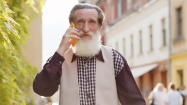 Yetişkin turist kıdemli adam arkadaşlarıyla uzaktan iletişim kuruyor. Yaşlı büyükbaba telefonda konuşuyor. Habersiz dedikodu. Şehir caddesinde yürüyor.