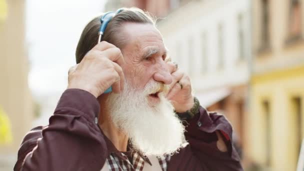 ワイヤレスヘッドフォンを着て 屋外で踊るお気に入りのエネルギッシュディスコロックNロールミュージックを聴くハッピーな老人 都市街道を歩く高齢のおじいちゃんの肖像画 — ストック動画