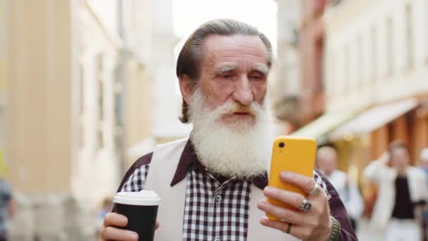 ハッピーシニア老人は インターネットソーシャルメディアWebアプリを閲覧するスマートフォンタイピングテキストメッセージを使用して 朝からコーヒーホットドリンクを楽しんでチャットします ストリートを歩く祖父の観光客 — ストック動画