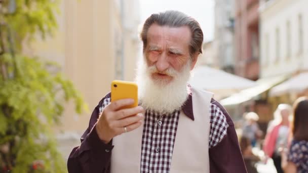 高齢者はスマートフォンのタイピングブラウジングを使用し 突然の宝くじの結果 悪いニュース 財産の損失 失敗に驚くようになります 高齢のおじいさんは 屋外の都市街道を歩いています タウンライフスタイル — ストック動画