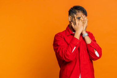 Meraklı Hintli adam elleriyle gözlerini kapatıyor ve parmaklarıyla casusluk yapıyor, saklanıyor ve dikizliyor, uzaklarda bir şeyler arıyor. Turuncu arka planda izole edilmiş Arap genç adam. Kopyalama alanı