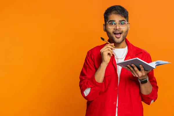 Excited happy journalist Indian man making notes, writing down thoughts with pen into notepad notebook diary, to do list good idea inspiration. Arabian guy isolated on orange background. Copy-space