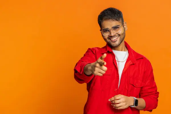 I choose you. Indian smiling man pointing finger to camera, looking with playful happy expression, making winner choice, showing direction. Arabian guy isolated on orange studio background. Copy-space