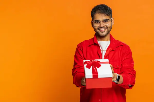Smiling Indian man presenting birthday gift box stretches out hands, offer wrapped present career bonus celebrating party promotion discount sale. Arabian guy isolated on orange background. Copy-space
