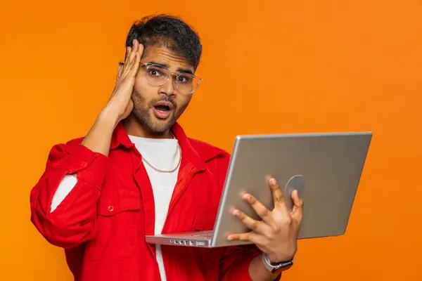 Displeased Indian man use laptop notebook typing browsing working, loses becoming surprised by lottery bad results fortune lost game fail computer virus, unlucky news. Arabian guy on orange background