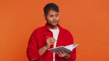 Düşünceli gazeteci genç Hintli adam notlar alıyor, not defteri günlüğüne kalemle düşünceler yazıyor, liste yapmak için, iyi fikir. Turuncu arka planda izole edilmiş Arap Hindu adam.