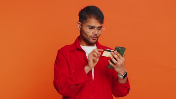 クレジットバンクカードとスマートフォンを使用して幸せな若いインド人男性は 支出を転送しながら オンラインショッピングを購入し 食料配達を注文します オレンジ色のスタジオの背景に孤立したアラビアの男 — ストック動画