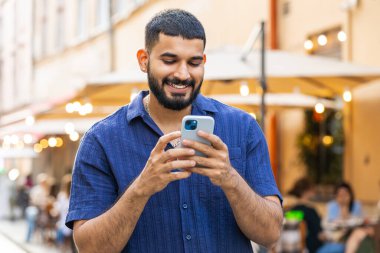 Mutlu, gülen, sakallı Hintli adam akıllı telefon kullanıyor, mesajlar yazıyor, internetteki sosyal medya uygulamalarını tarıyor açık havada sohbet ediyor. Şehir caddesinde yürüyen bir turist. Kasaba yaşam tarzları