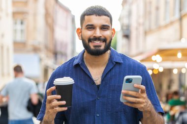 Mutlu Hintli adam akıllı telefon kullanarak mesajlar yazıyor internette sosyal medya uygulamalarını tarıyor internette sohbet ediyor ve sabahları dışarıda sıcak kahve içip eğleniyor. Şehir caddesinde yürüyen bir adam.
