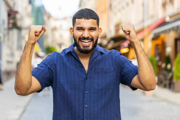 Bearded young adult man shouting, celebrating success, winning, goal achievement, good news, lottery jackpot luck, victory outdoors. Indian guy walking in urban city sunshine street. Town lifestyles