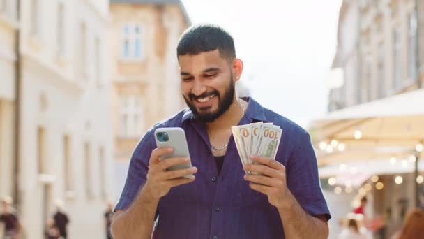 快乐富有的印度男人数着钞票 使用智能手机计算器应用程序 计划在网上订购礼物和食品 预订酒店房间 大胡子的游客在城市街道上 — 图库视频影像
