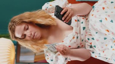 Aile bütçesi planlamak. Gülümseyen kızıl saçlı kadın nakit para sayıyor, akıllı telefon kullanıyor ev faturalarını hesaplıyor. Gelirinden tatmin olan ve planlı tatil ve hediyeler için para biriktiren bir kız. Dikey