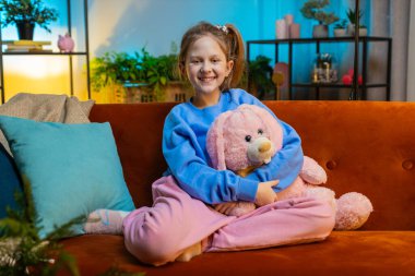 10 yaşındaki mutlu, beyaz, liseli bir kızın yakın plan portresi. Evdeki oyun odasında turuncu koltukta oturan sevimli çocuk kameraya bakıyor. At kuyruğu saç stili.