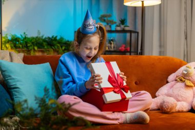 Mutlu küçük kız doğum günü şapkalı şapka hediye paketi takıyor ve kurdele takıyor. Ergen bayan çocuk parti olayını kutluyor, doğum günü hediyesini kanepede açıyor.